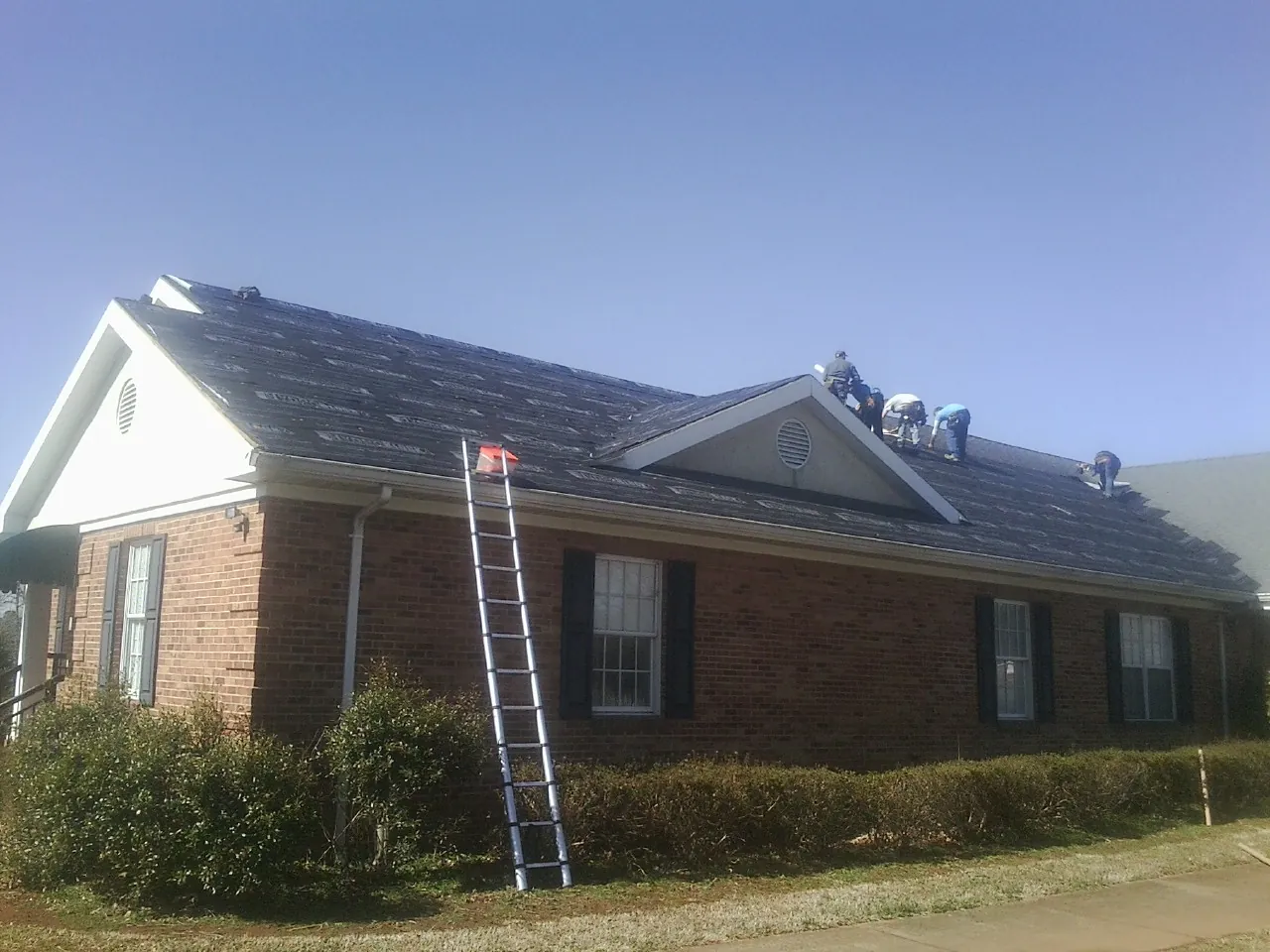 Crew tearing shingles off a brick ranch roof with a ladder set — Emergency Roof Repair work by our crew in Lindsey