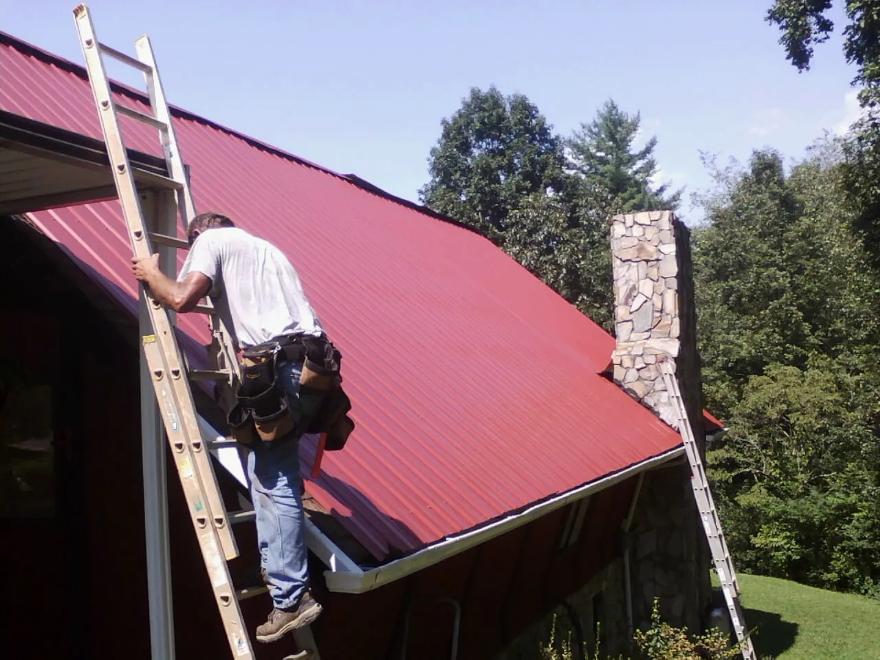 Roofer on a ladder beside a red metal roof and stone chimney — Soffit & Fascia Repair by Roofing Brothers in Lindsey