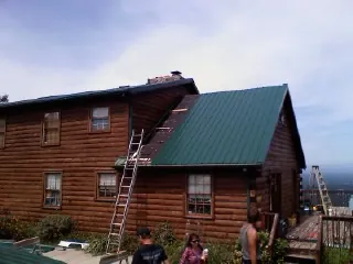 Green metal roof going on a two story log home with ladders set in Lindsey